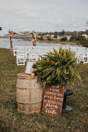 Rustic wooden barrels with greenery and wooden wedding signs arranged on grass for an outdoor ceremony setup.