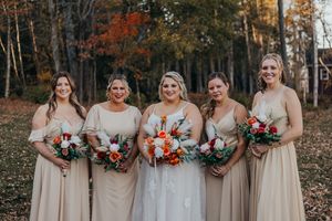Wedding party in beige dresses holds colorful autumn bouquets with orange and burgundy flowers in a fall forest setting.