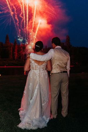 A couple watches vibrant red fireworks illuminate the night sky during their outdoor wedding celebration.
