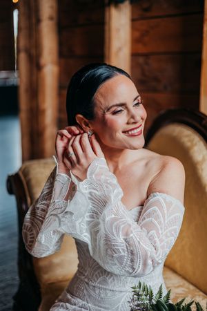 Bride wearing elegant lace off-shoulder wedding dress smiles while putting on earrings in rustic wooden interior.