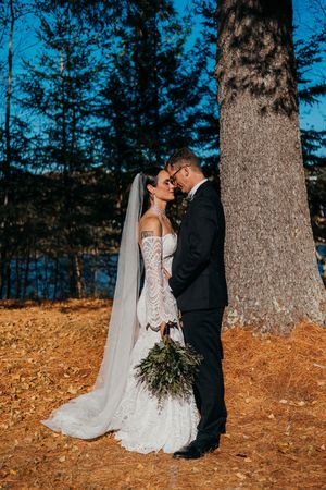 A romantic wedding couple poses against a large tree trunk with autumn leaves scattered on the ground.
