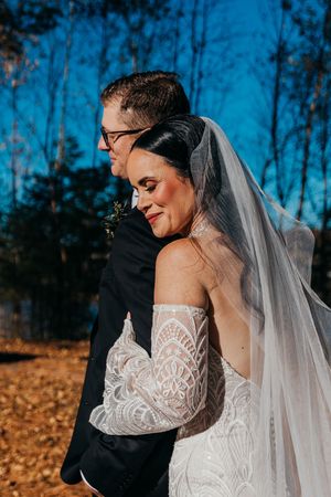 A bride in a lace off-shoulder wedding dress and veil smiles while embracing her groom outdoors on an autumn day.