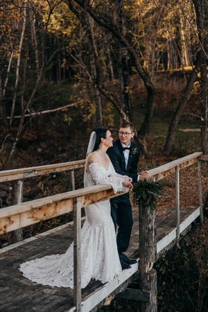 A couple in wedding attire share an intimate moment on a rustic wooden bridge surrounded by autumn trees.