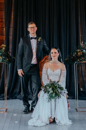 A wedding party in formal attire poses against a dark backdrop with elegant floral arrangements and greenery.