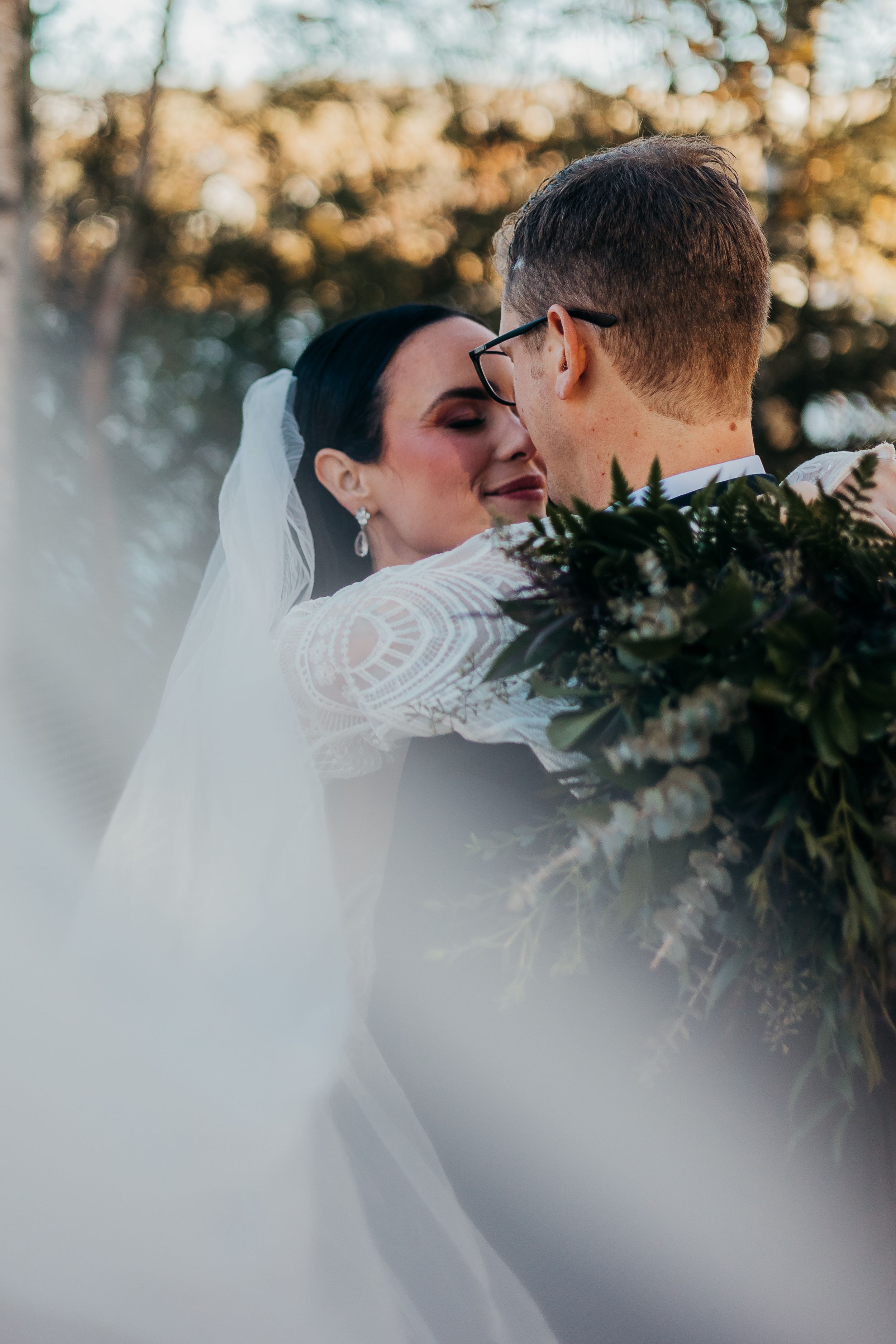 A romantic wedding moment captured through evergreen branches during golden hour.