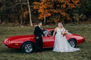 Newlywed couple poses with classic red vintage car in autumn outdoor wedding photoshoot.