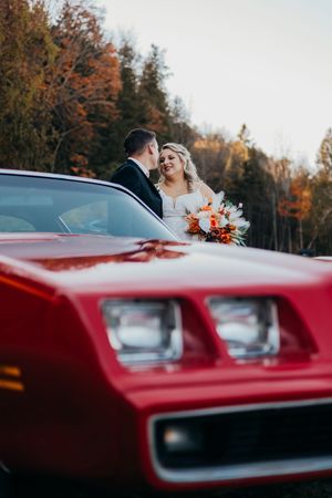 A couple stands behind a vintage red car during an autumn wedding photoshoot with colorful fall foliage in background.