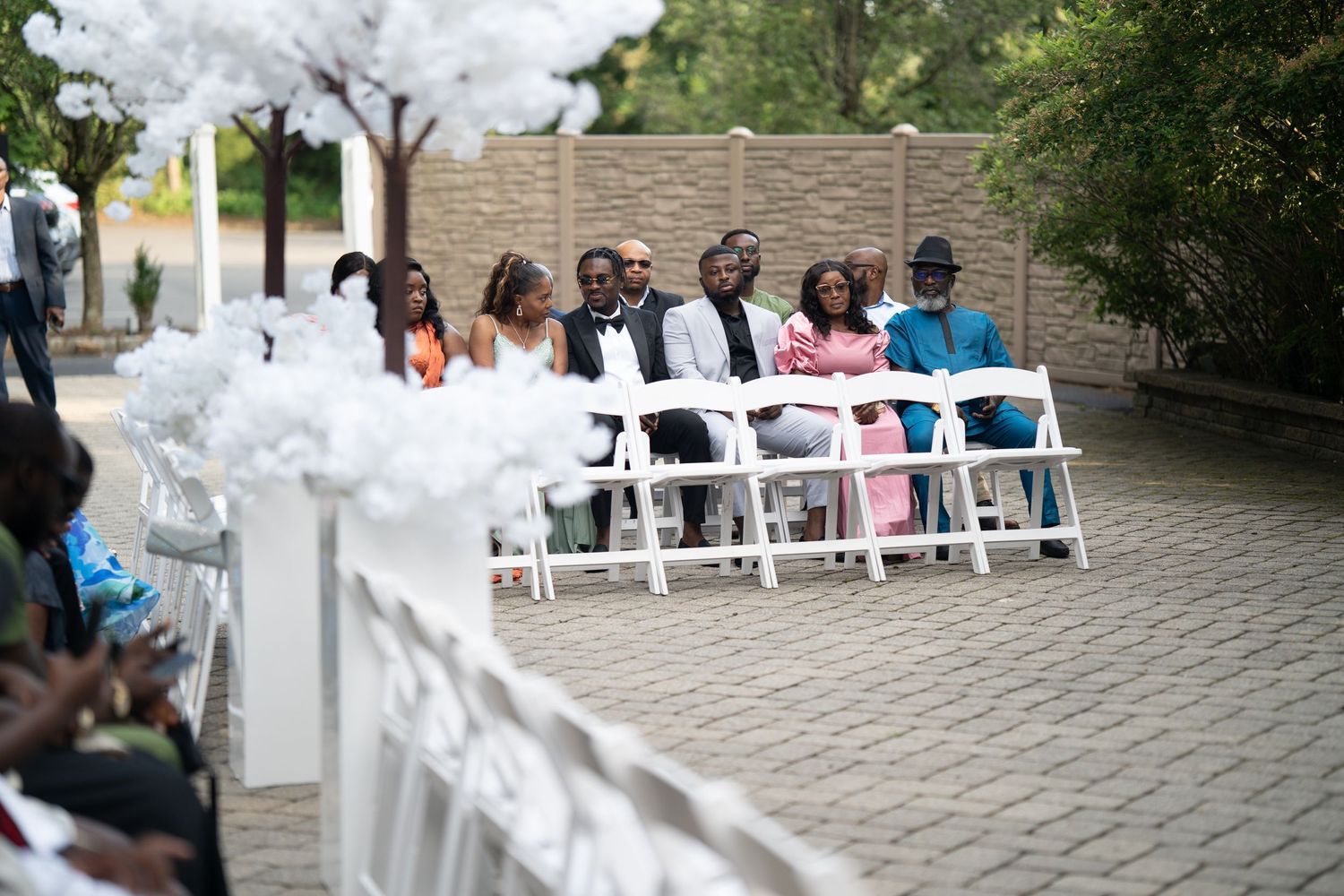 Young ring bearer in white tuxedo jacket and black pants stands on curved stone path during outdoor wedding ceremony.