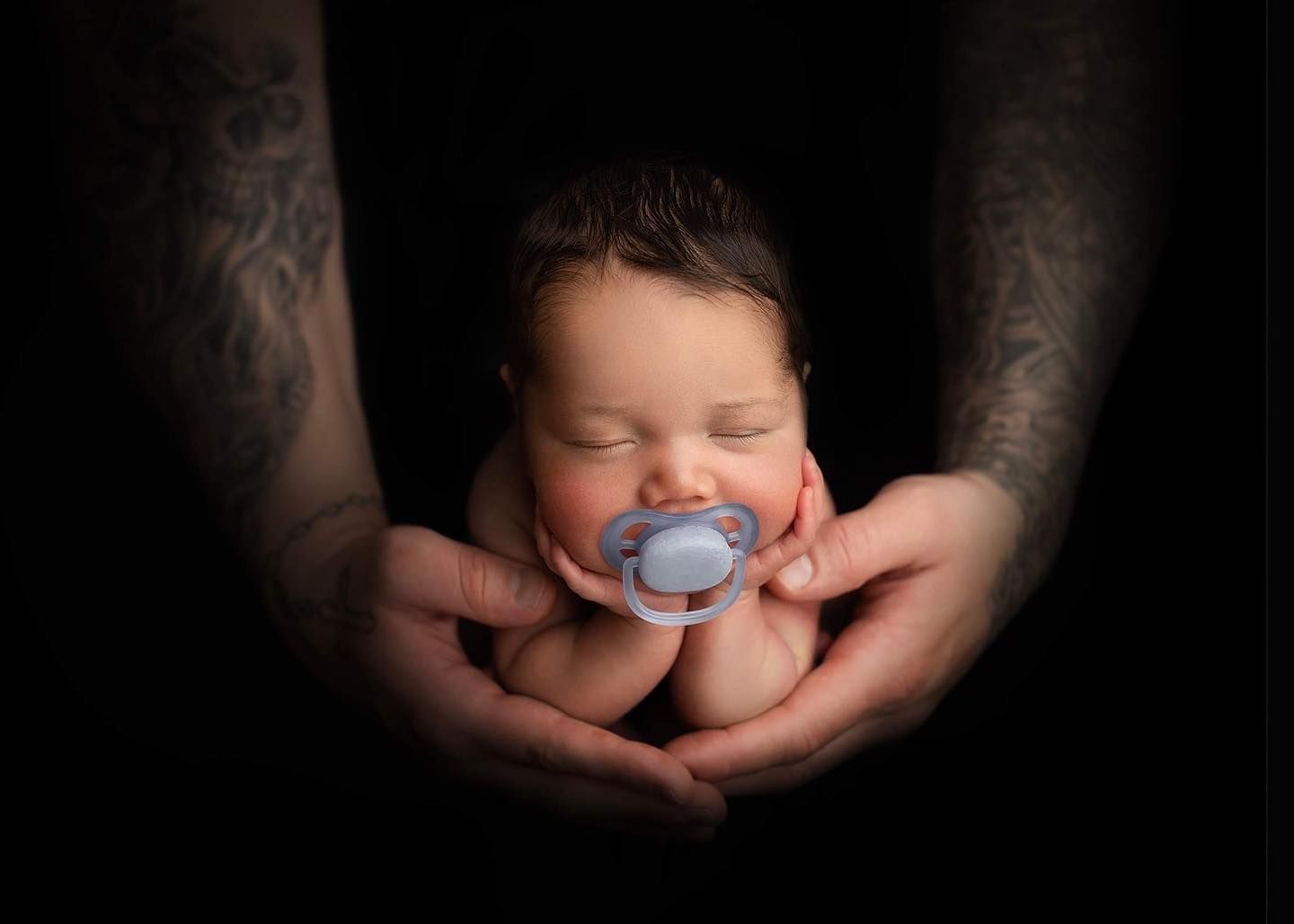 A sleeping newborn cradled in tattooed hands with a blue pacifier against a dark background.