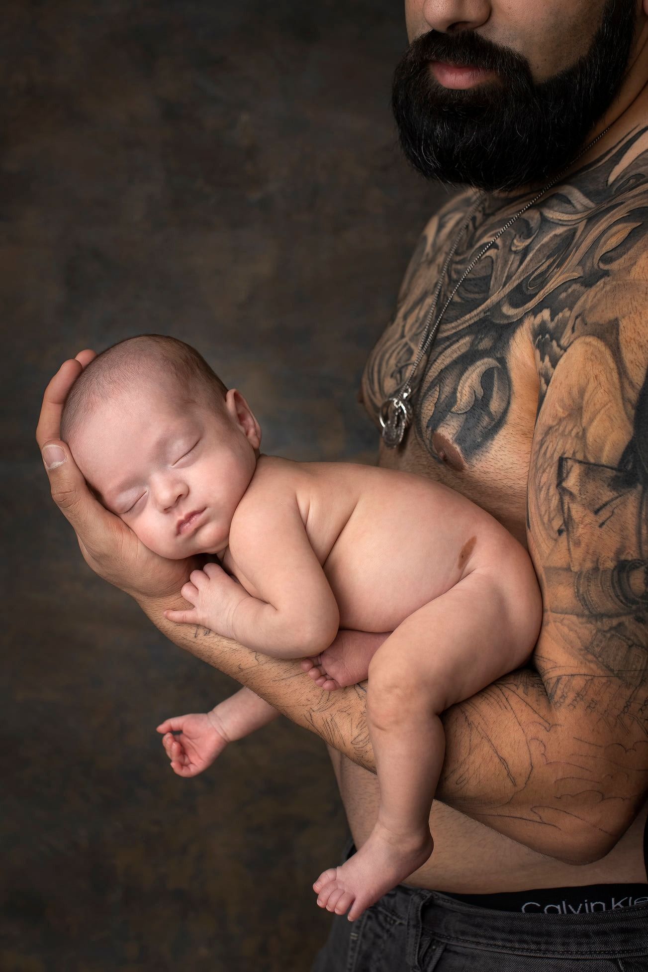 A sleeping newborn baby nestled in tattooed arms against a dark studio backdrop.