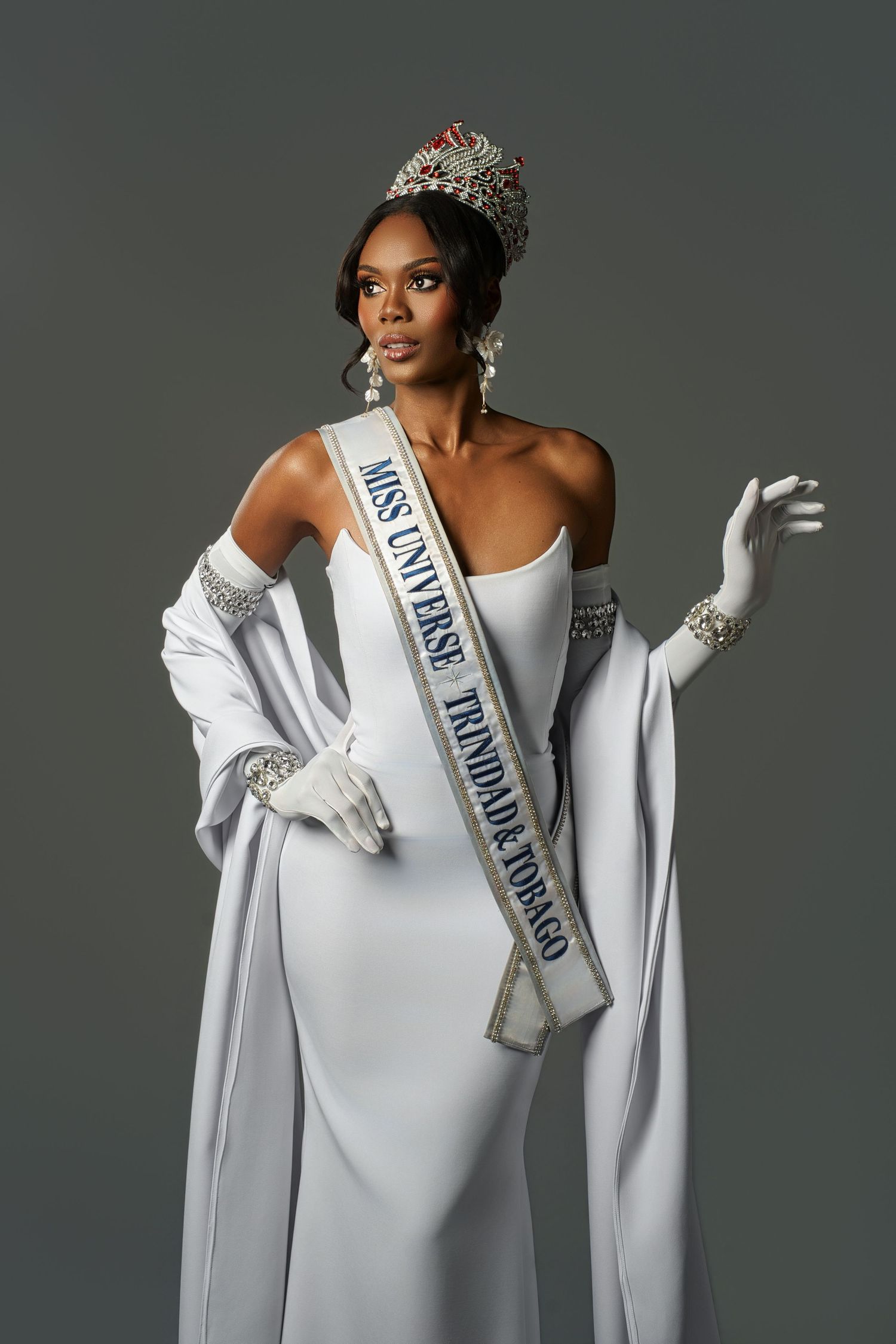 Beauty pageant contestant wearing a white off-shoulder gown with crystal details and an official sash in a professional photoshoot.