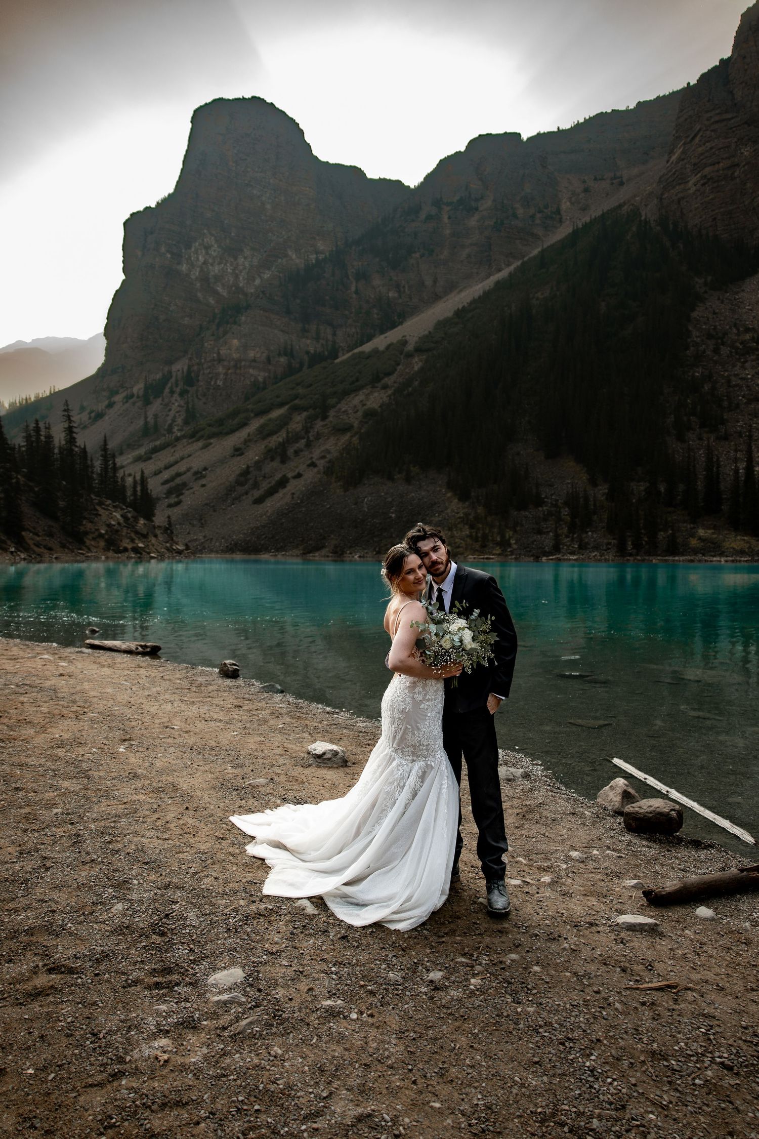 A couple shares an intimate moment on the rocky shore of a turquoise mountain lake surrounded by dramatic peaks.