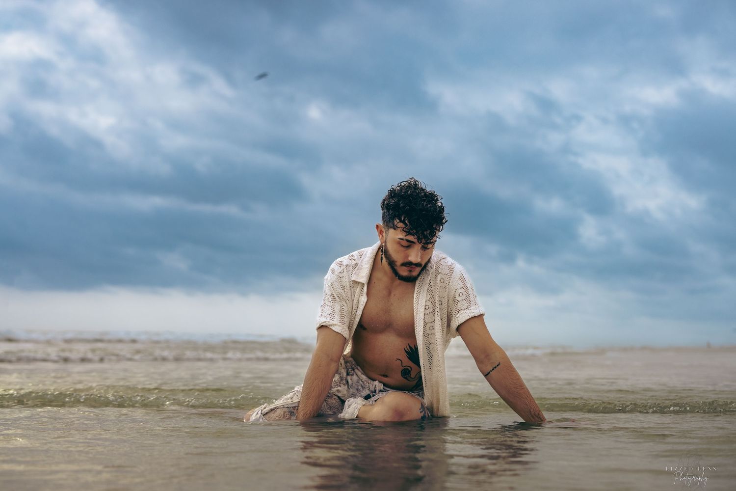 Shirtless man kneeling in ocean waves under dramatic cloudy sky at beach.