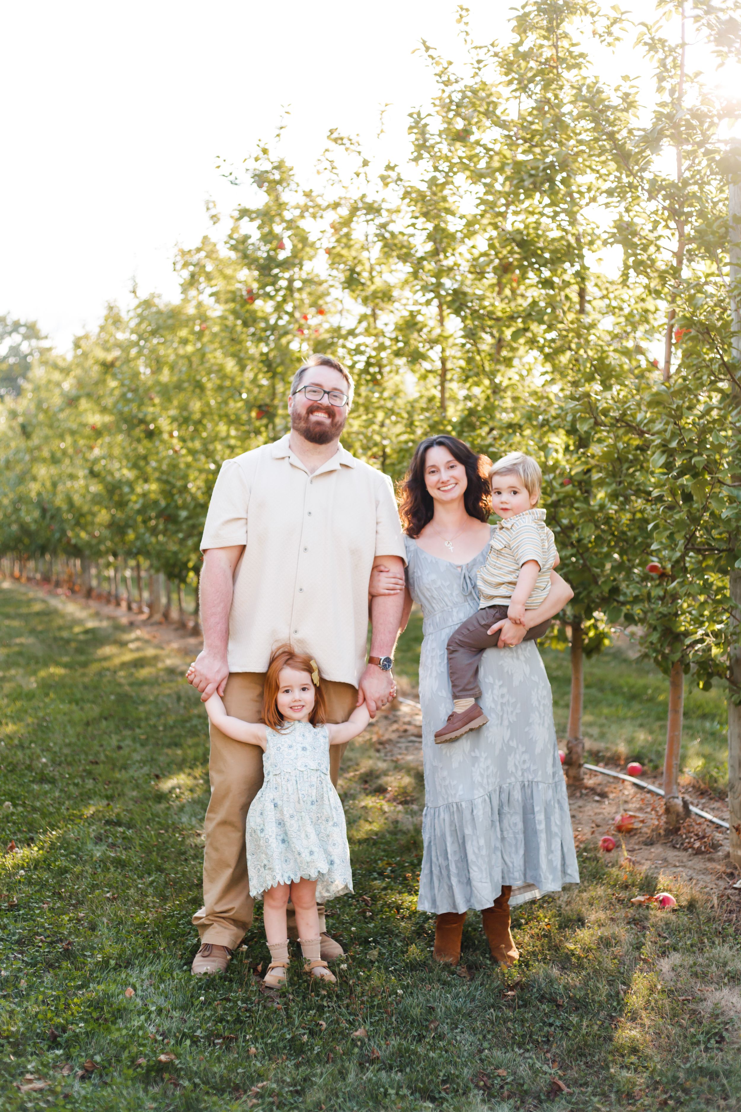A family poses together for a portrait in front of rows of apple trees at an orchard during golden hour.
