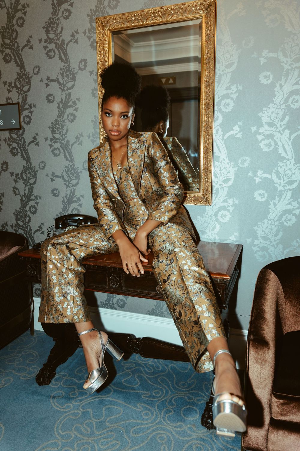 Fashion editorial featuring metallic gold patterned suit against floral wallpaper with ornate gold frame.