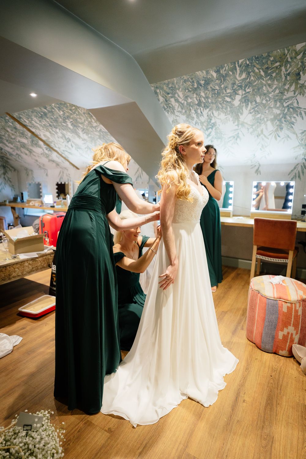 Bridesmaids in dark green dresses helping bride with wedding gown preparations.