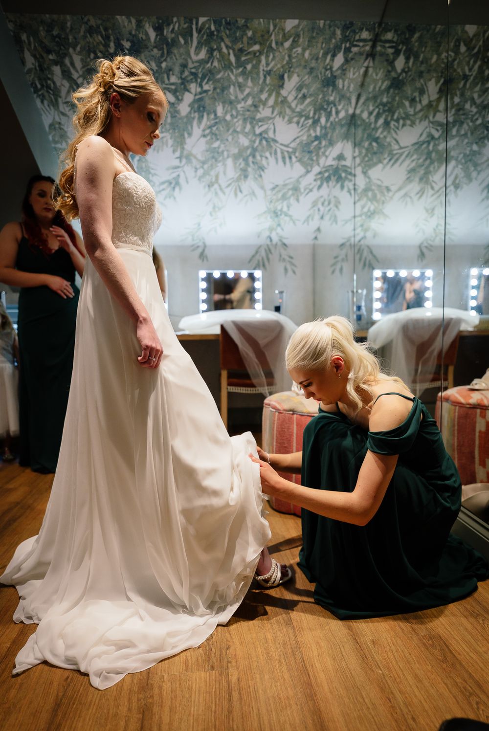 Wedding dress preparation scene in a dressing room with bridesmaids helping with final touches.
