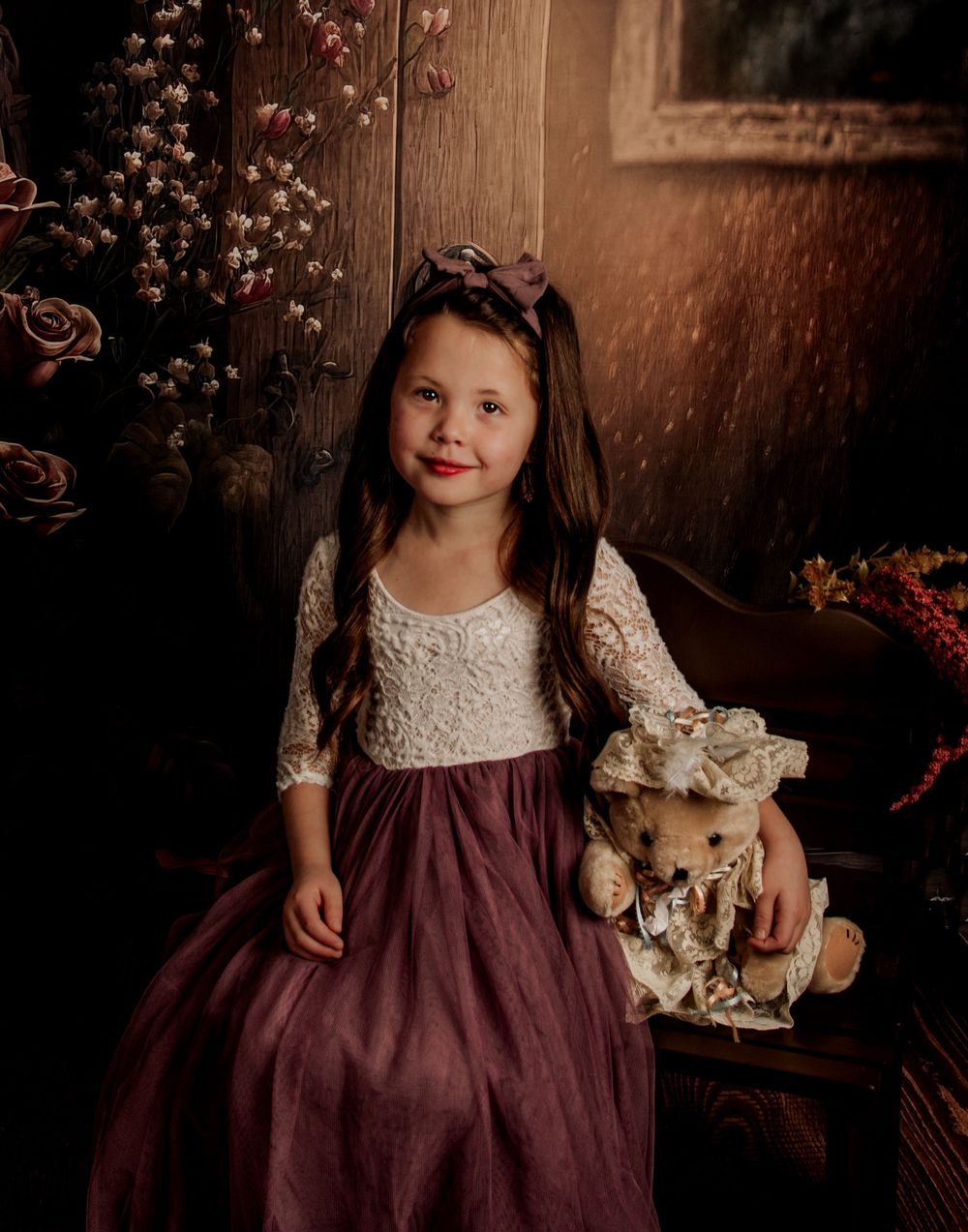 A child in a burgundy dress with lace details poses with a plush toy against a rustic wooden backdrop.
