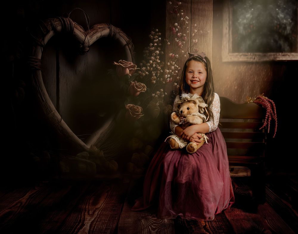 A dreamy portrait in dark tones features a child in vintage dress holding a teddy bear beside a heart-shaped window.