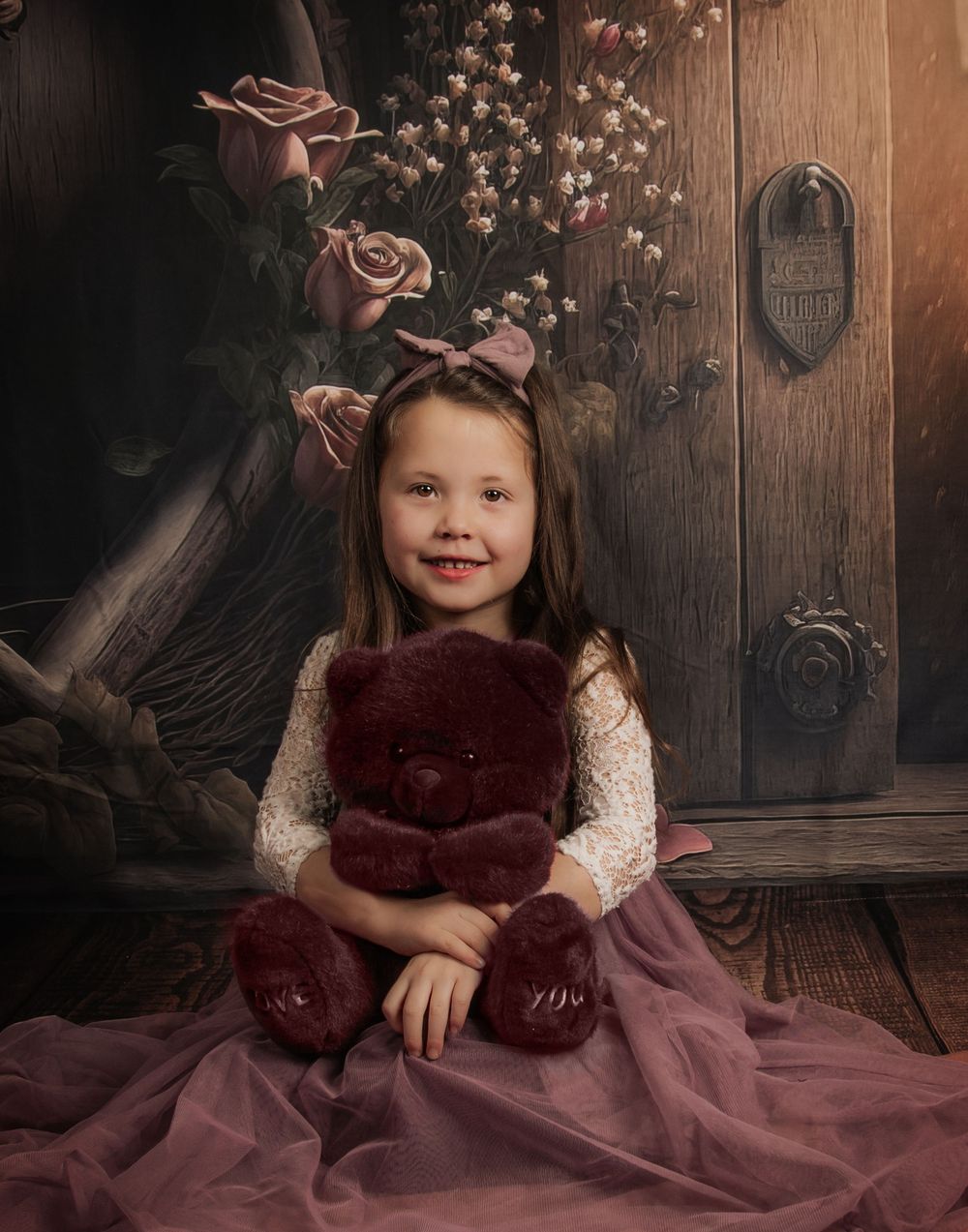 A whimsical portrait featuring a burgundy dress, teddy bear, and vintage wooden door with floral accents.