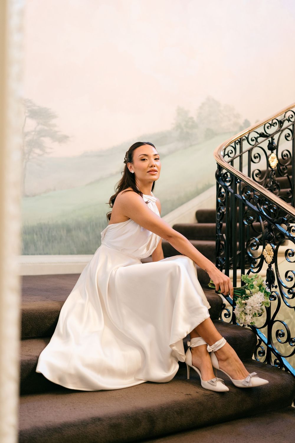 A bride in a flowing white dress reclines elegantly on dark stairs against an ornate railing with mountains in the background.