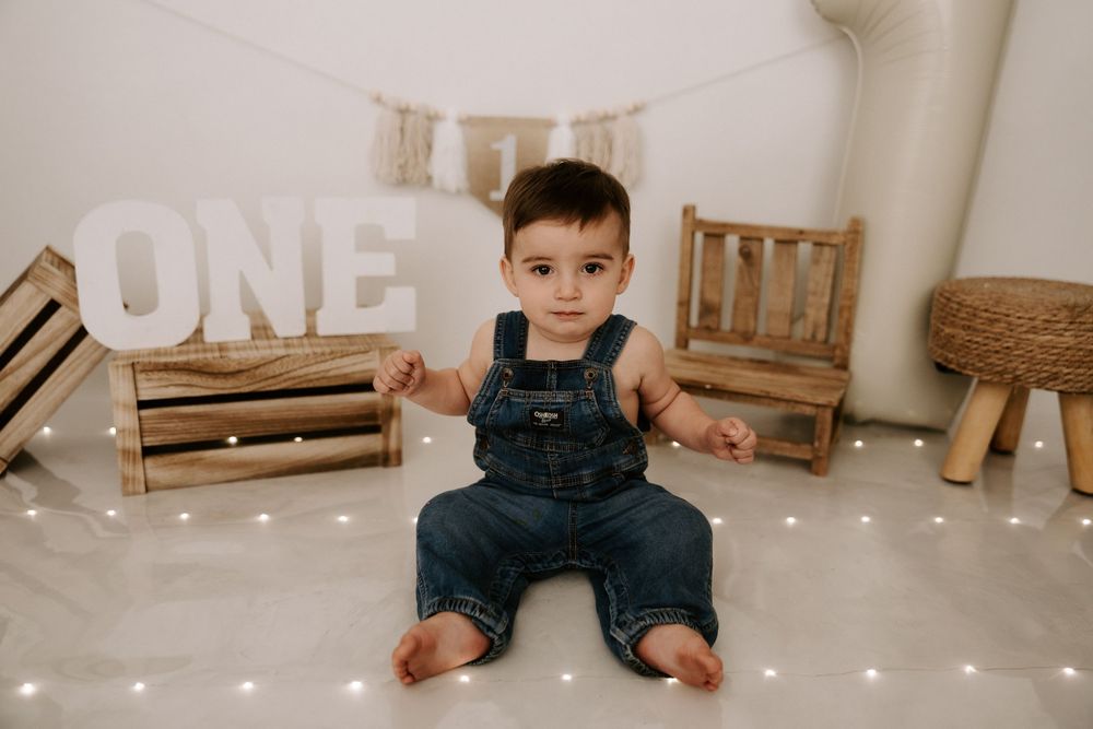 A toddler in denim overalls sits on the floor with wooden crates and 'ONE' letters in a rustic styled photoshoot setup.