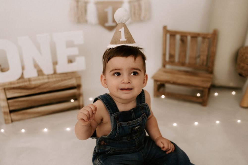 A small child in denim overalls sits on the floor with a party hat, smiling next to wooden decor and lights.