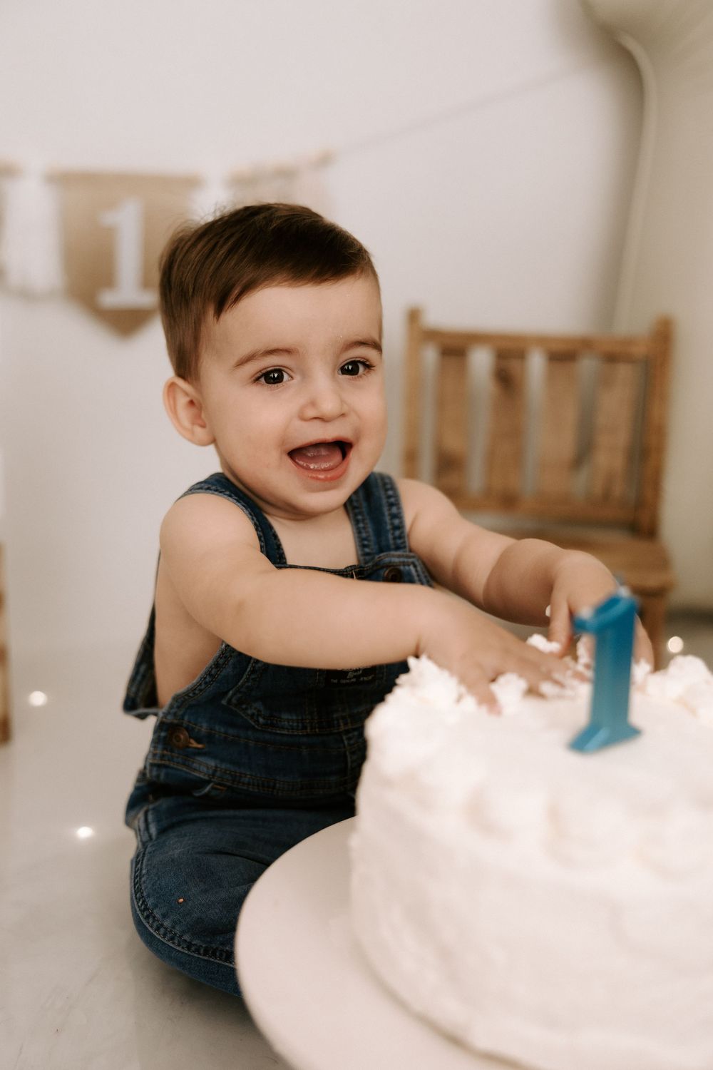 A happy baby in denim overalls plays with white frosting or cream during a cake smash photo session.