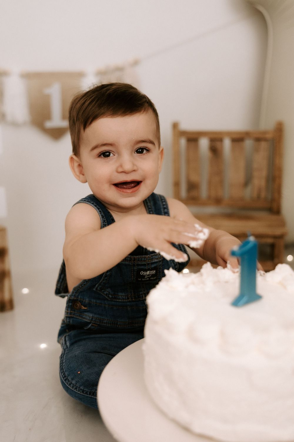 A happy toddler in denim overalls reaches for a white birthday cake with a blue number one candle on top.