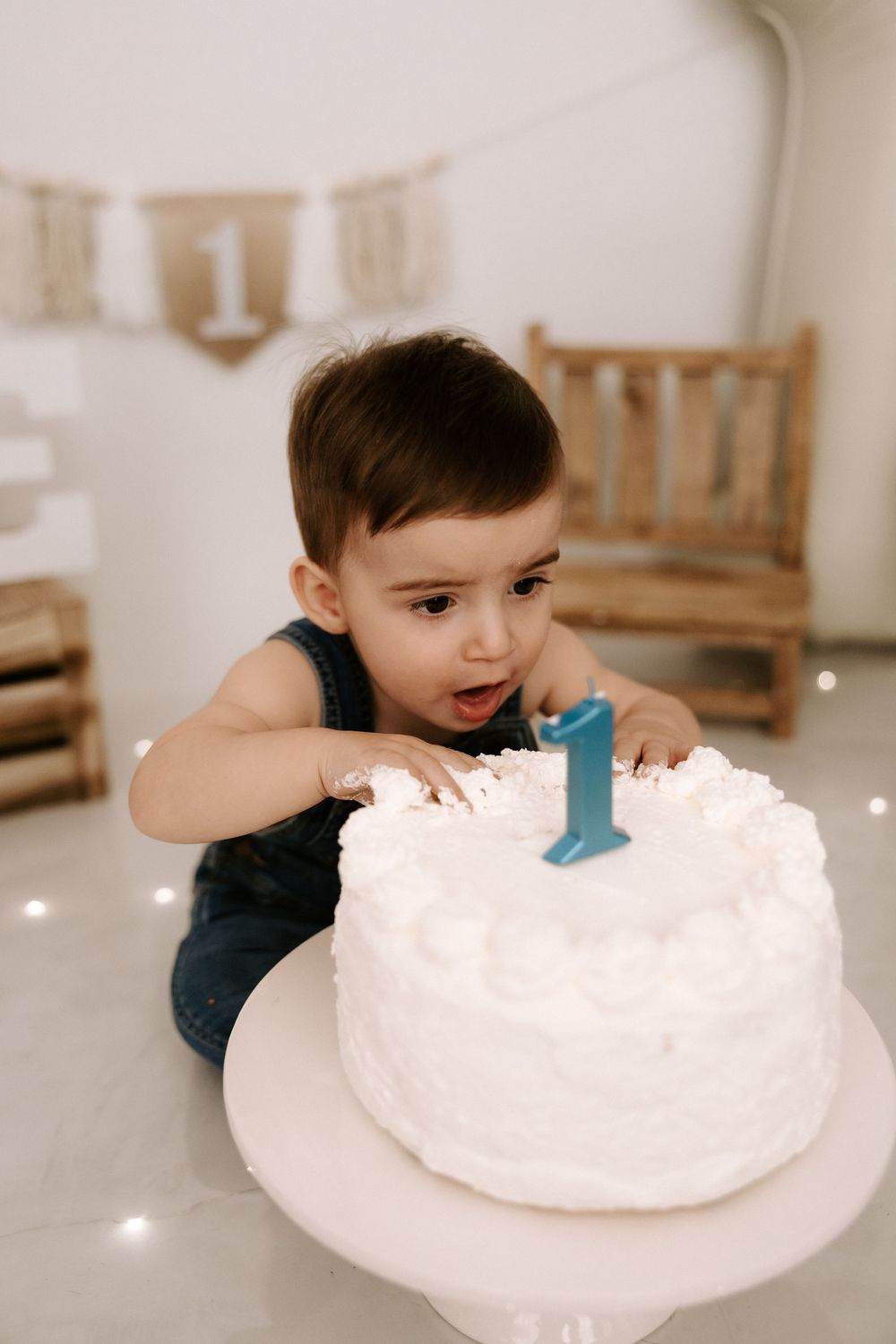A small child in dark overalls reaches for a white birthday cake with a blue number one candle on top.