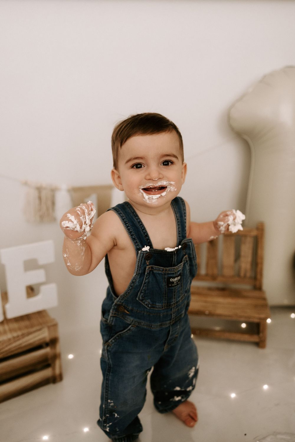A joyful toddler in denim overalls plays and laughs while sitting on a light colored floor.