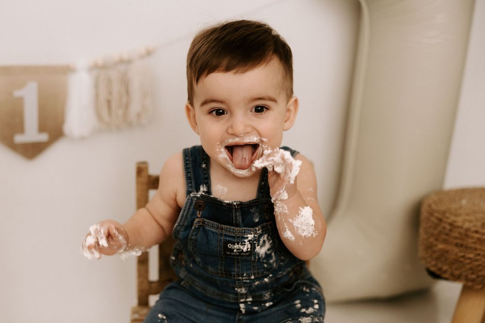 A joyful toddler in denim overalls plays with cake frosting during a first birthday photoshoot.