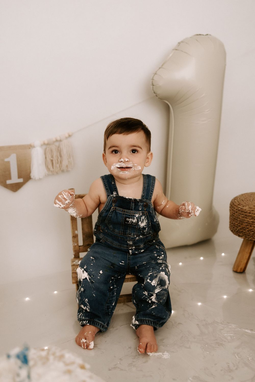 A baby in denim overalls sits on a white floor during a cake smash photo session with sparkly decorations.