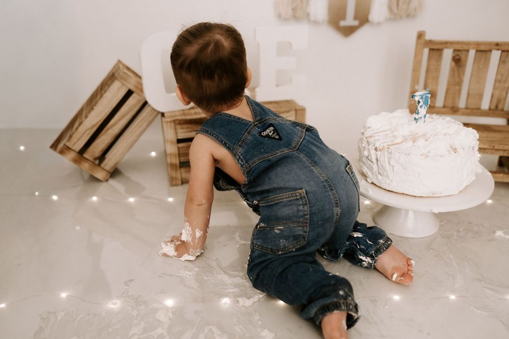 A small child in overalls crawls on a messy floor near a white birthday cake with scattered decorations and chairs.
