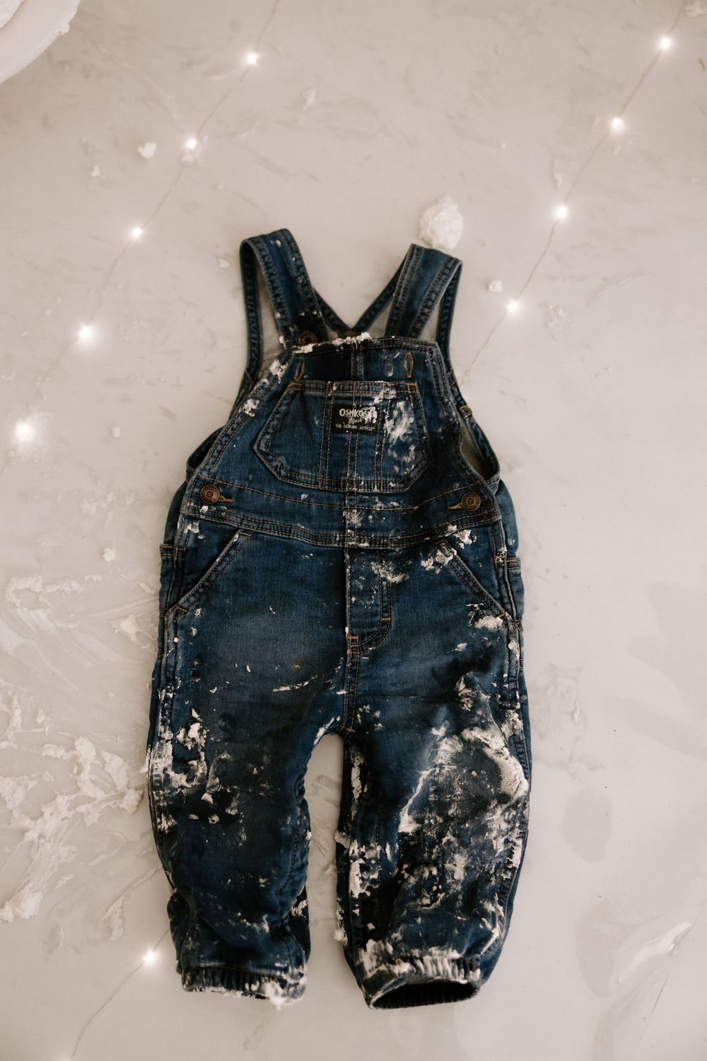 Messy denim baby overalls covered in white frosting and cake stains lying flat on a light surface.