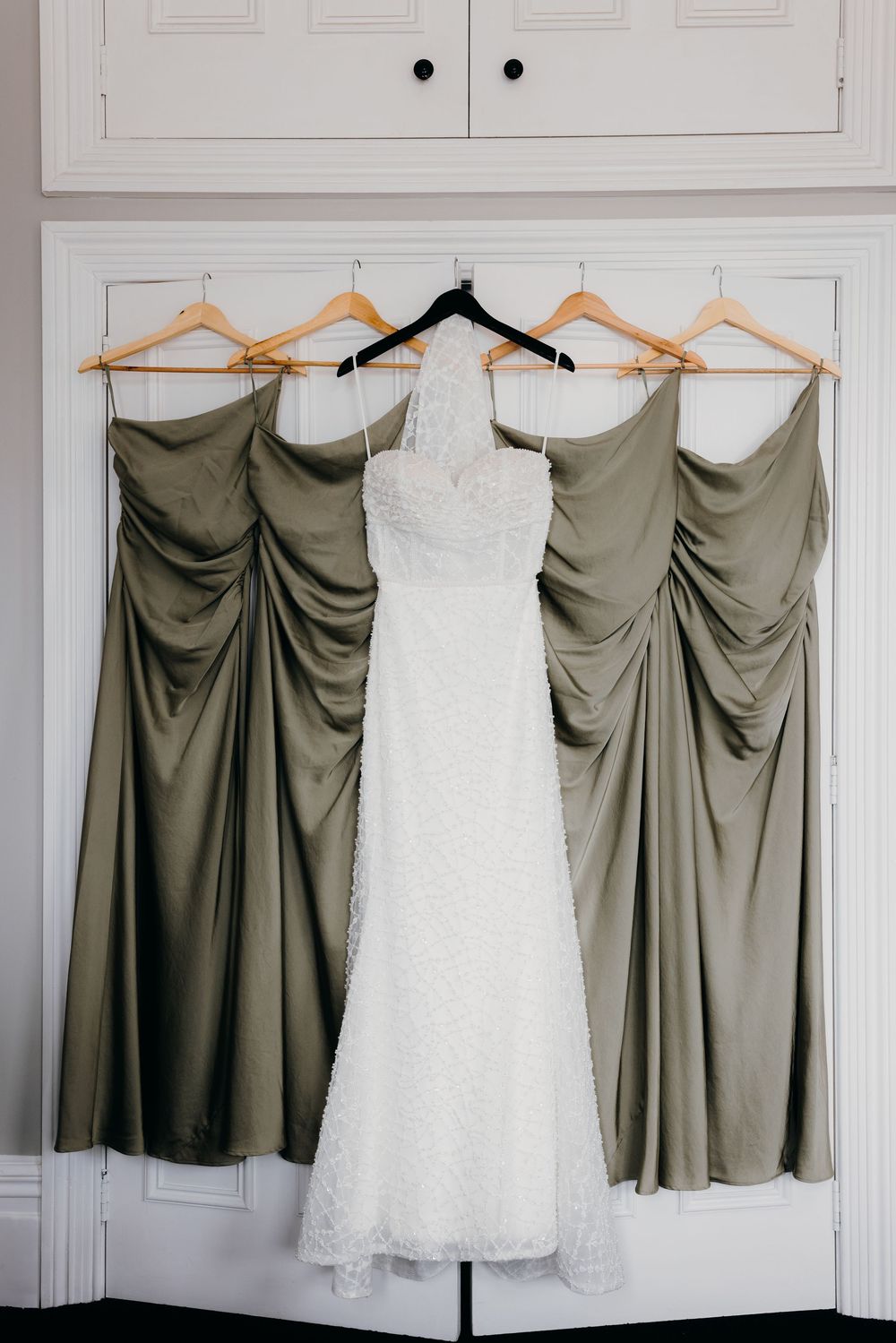 A white lace wedding dress hangs between sage green bridesmaid dresses on wooden hangers against a white wall.