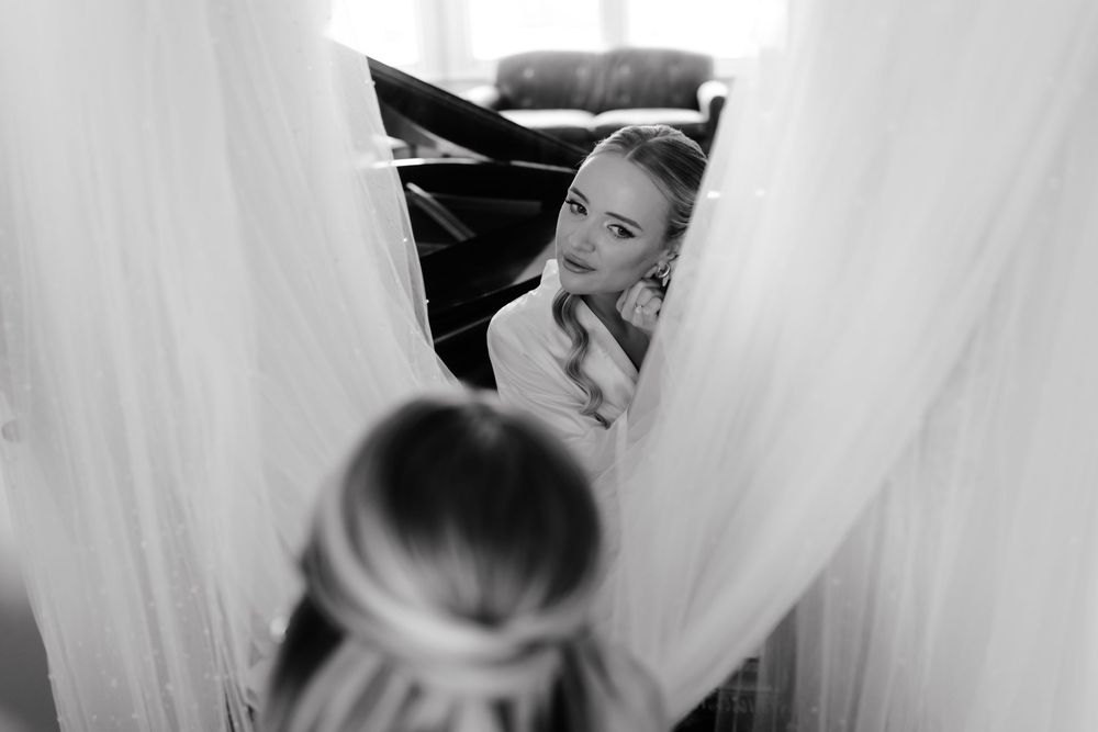 A black and white reflection in a mirror shows someone peeking through white curtains or drapes.