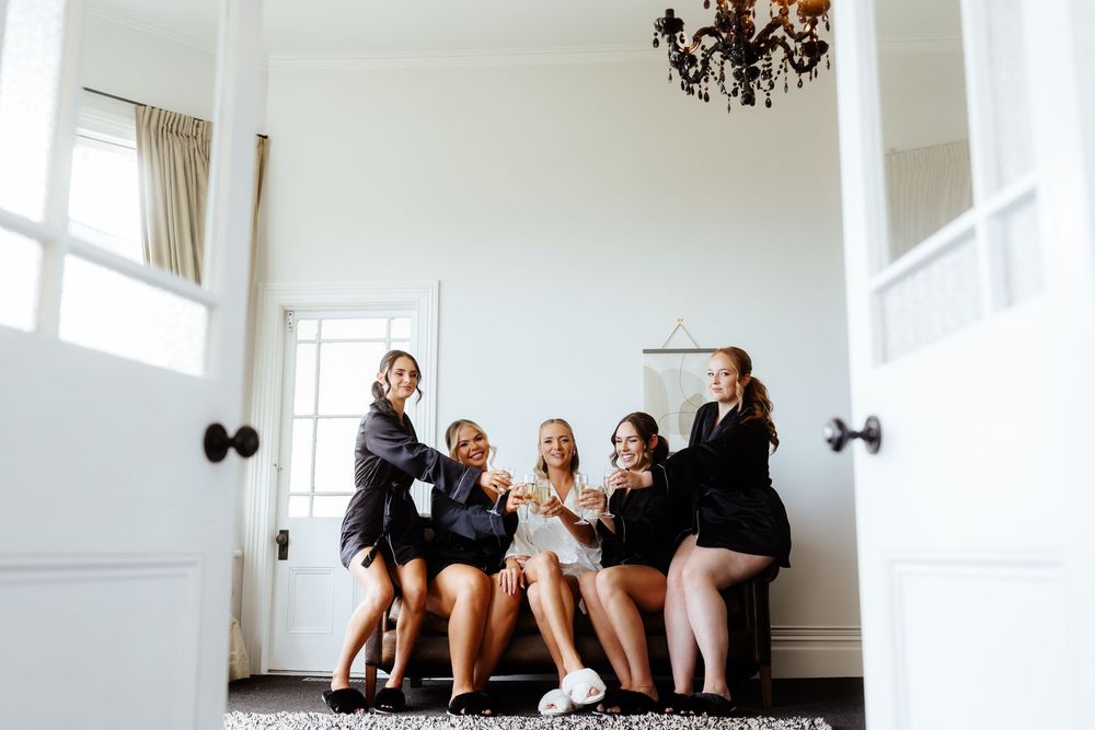 A group of dancers in black outfits sit in a bright white room with hardwood floors and natural light.