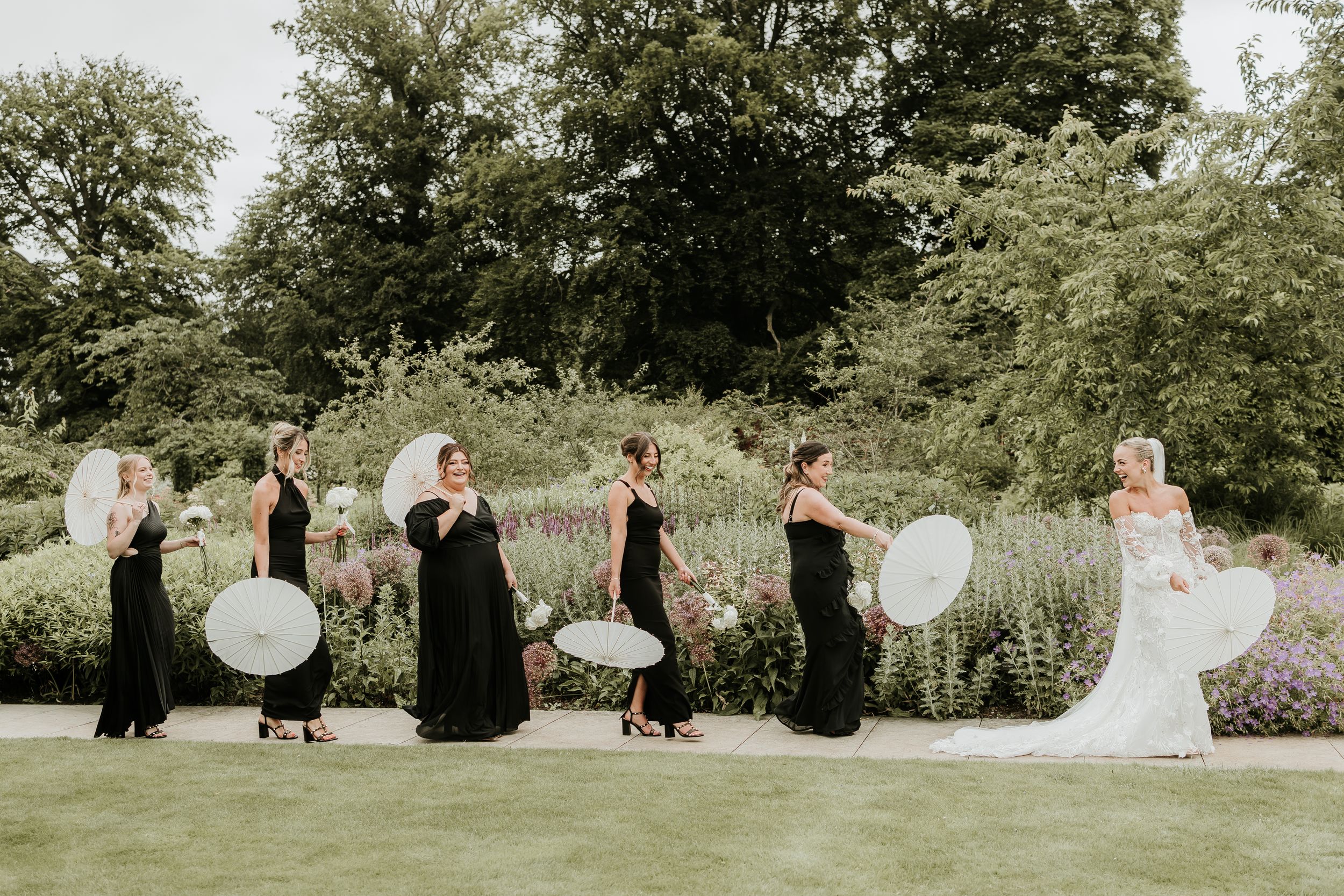 A bride and bridesmaids in black dresses walk in a line holding white balloons in the gardens of the fig house at Middleton lodge.