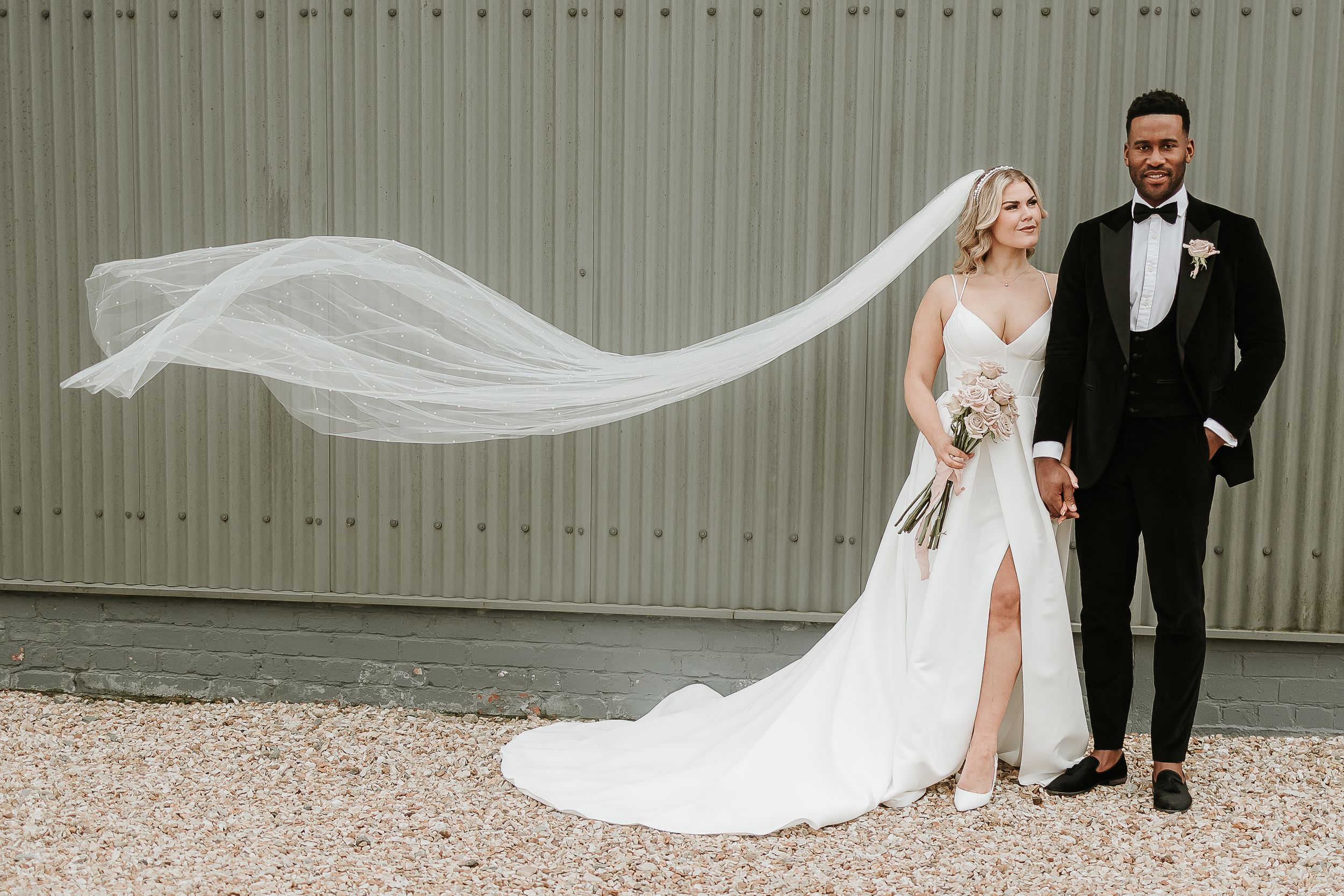 A flowing white wedding veil billows in the wind against a light grey wall as a couple stands together. Abbey farm wedding venue.