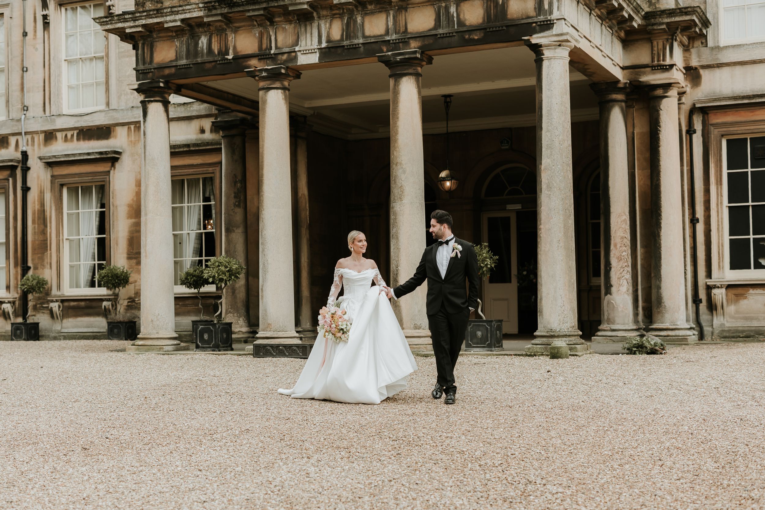 A romantic wedding couple poses in front of an elegant columned building with gravel path.