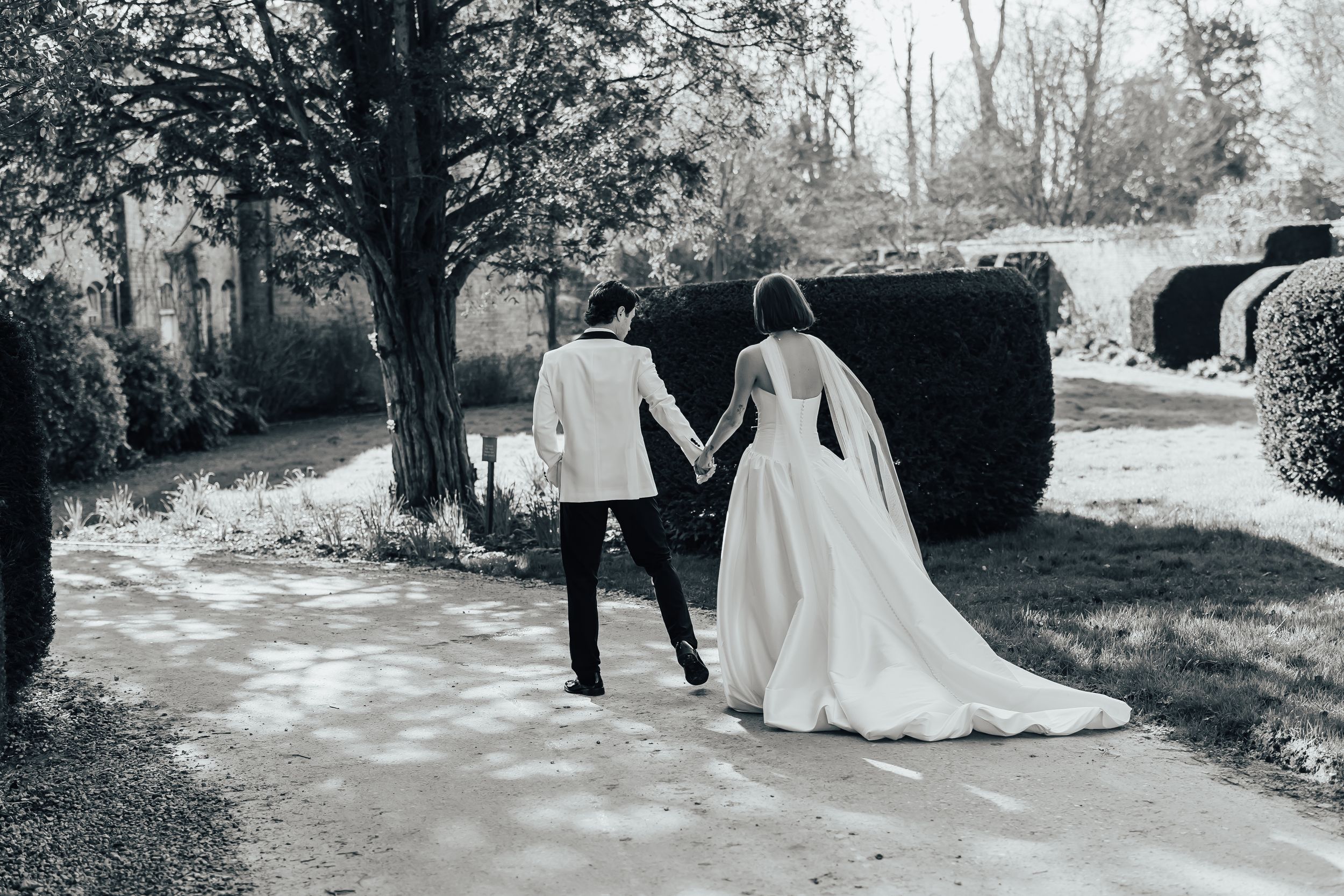 A black and white wedding couple walks along a scenic tree-lined path in a romantic outdoor setting.