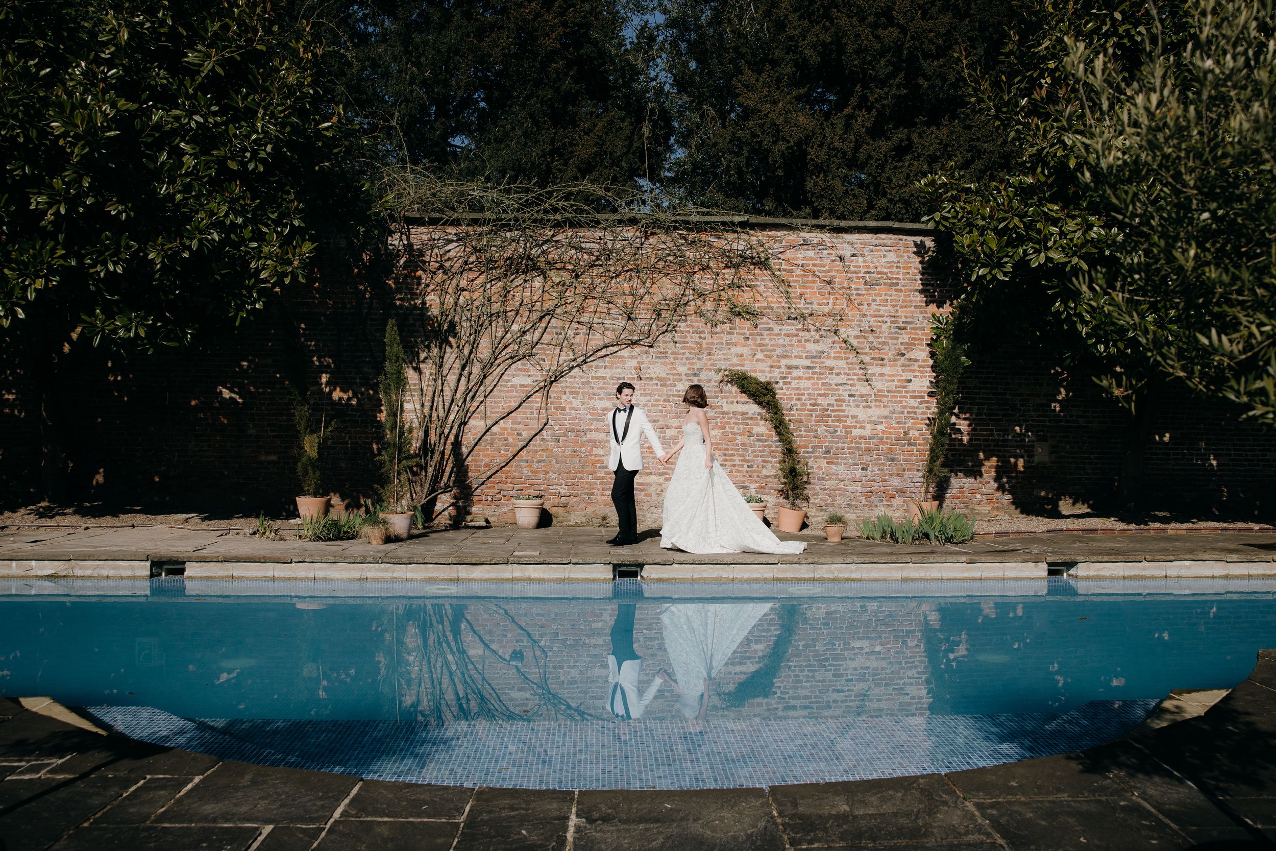 Elegant couple stands beside a serene blue swimming pool against a rustic stone wall in a garden setting.