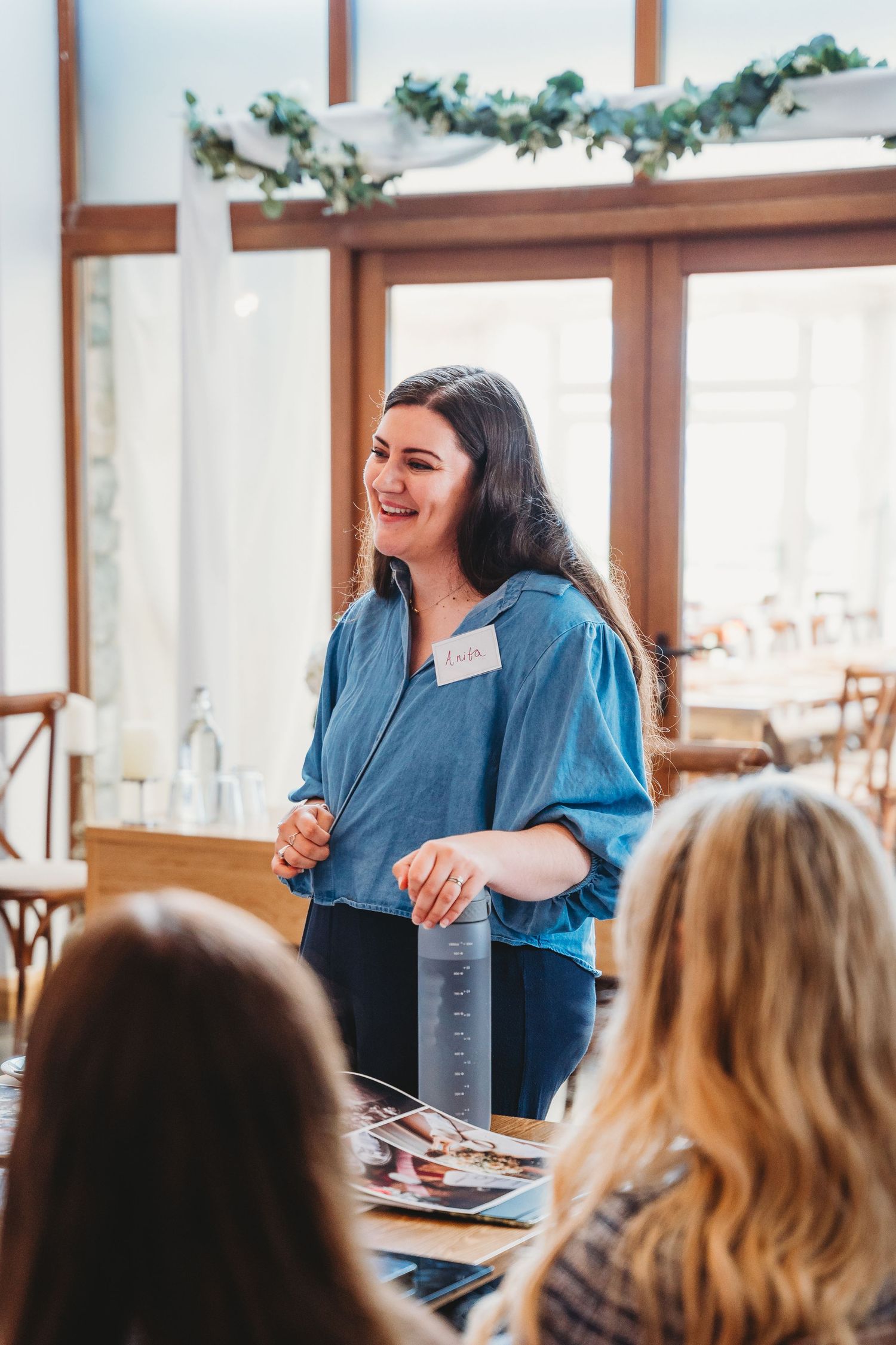 A speaker in a blue shirt engages with an attentive audience during a presentation in a bright, window-lit room.