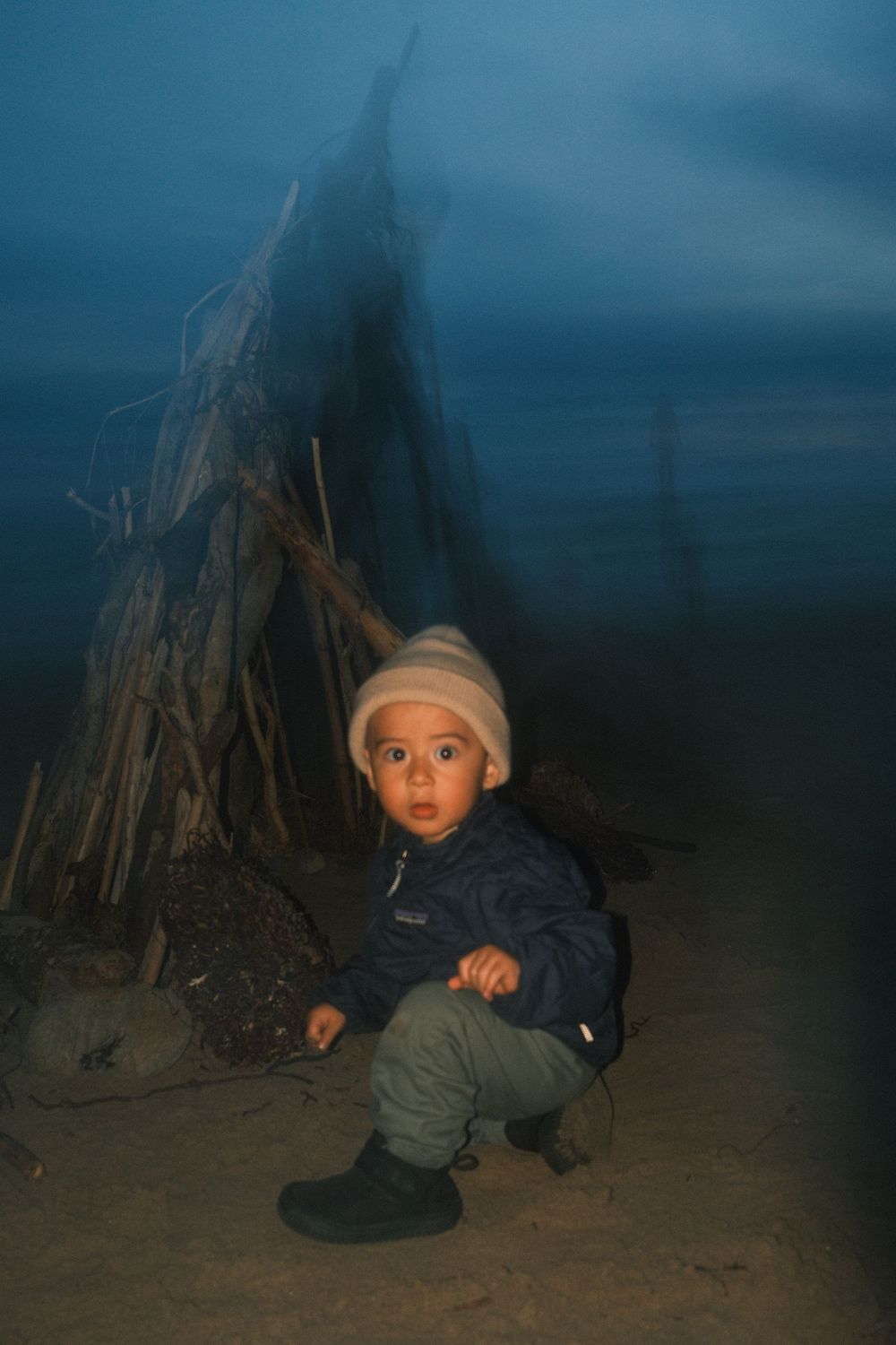 A small child wearing a winter hat and coat crouches near a wooden teepee structure at dusk.