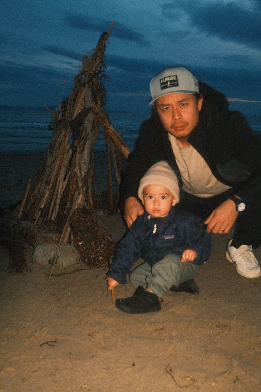 Family enjoying evening beach bonfire with driftwood teepee structure on sandy shore at dusk.