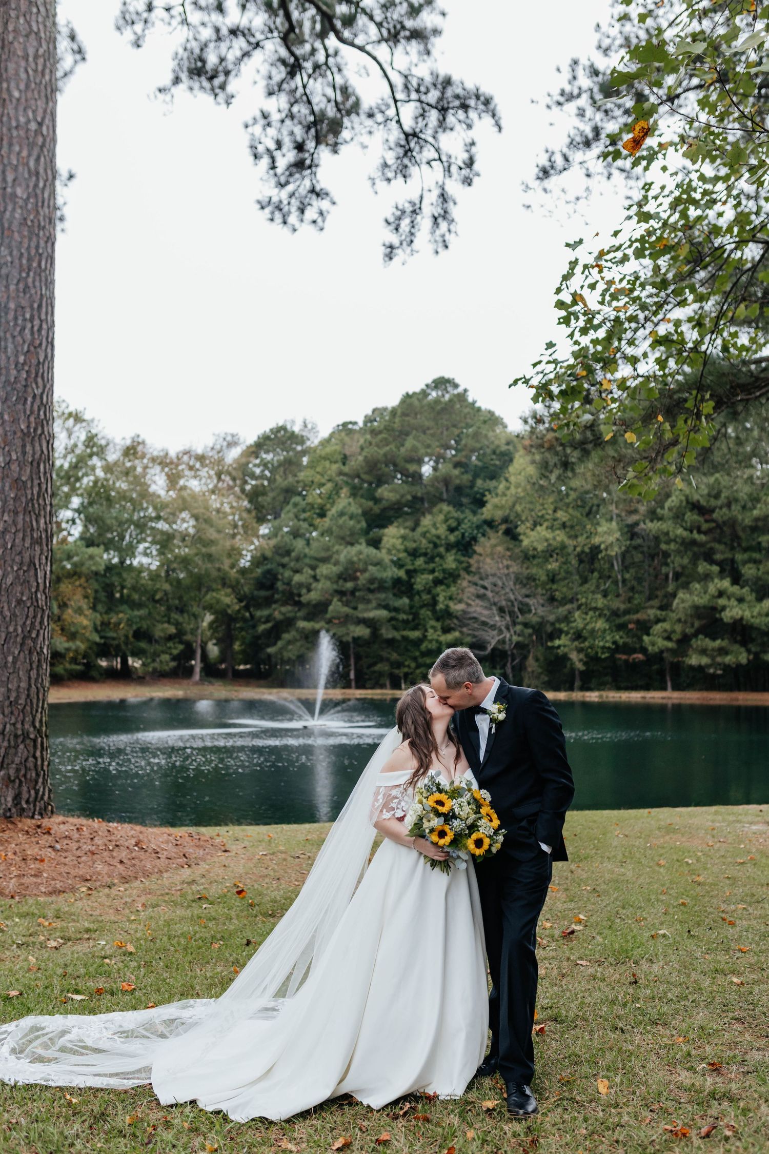 A couple in wedding attire stands together on a grassy lawn near a lake with a fountain in the background.