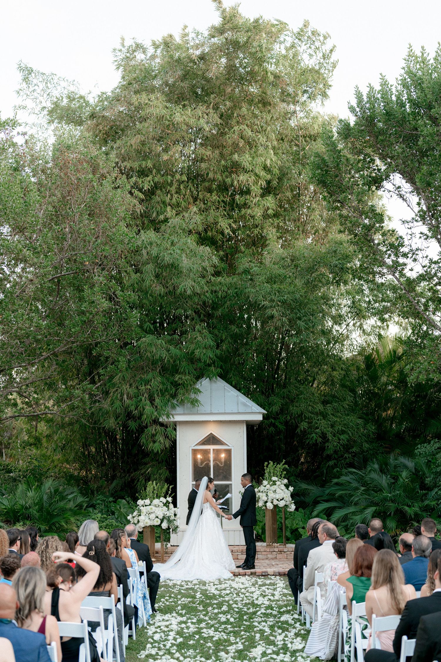 A bride in a flowing white wedding gown and veil poses outside a rustic building under oak trees.