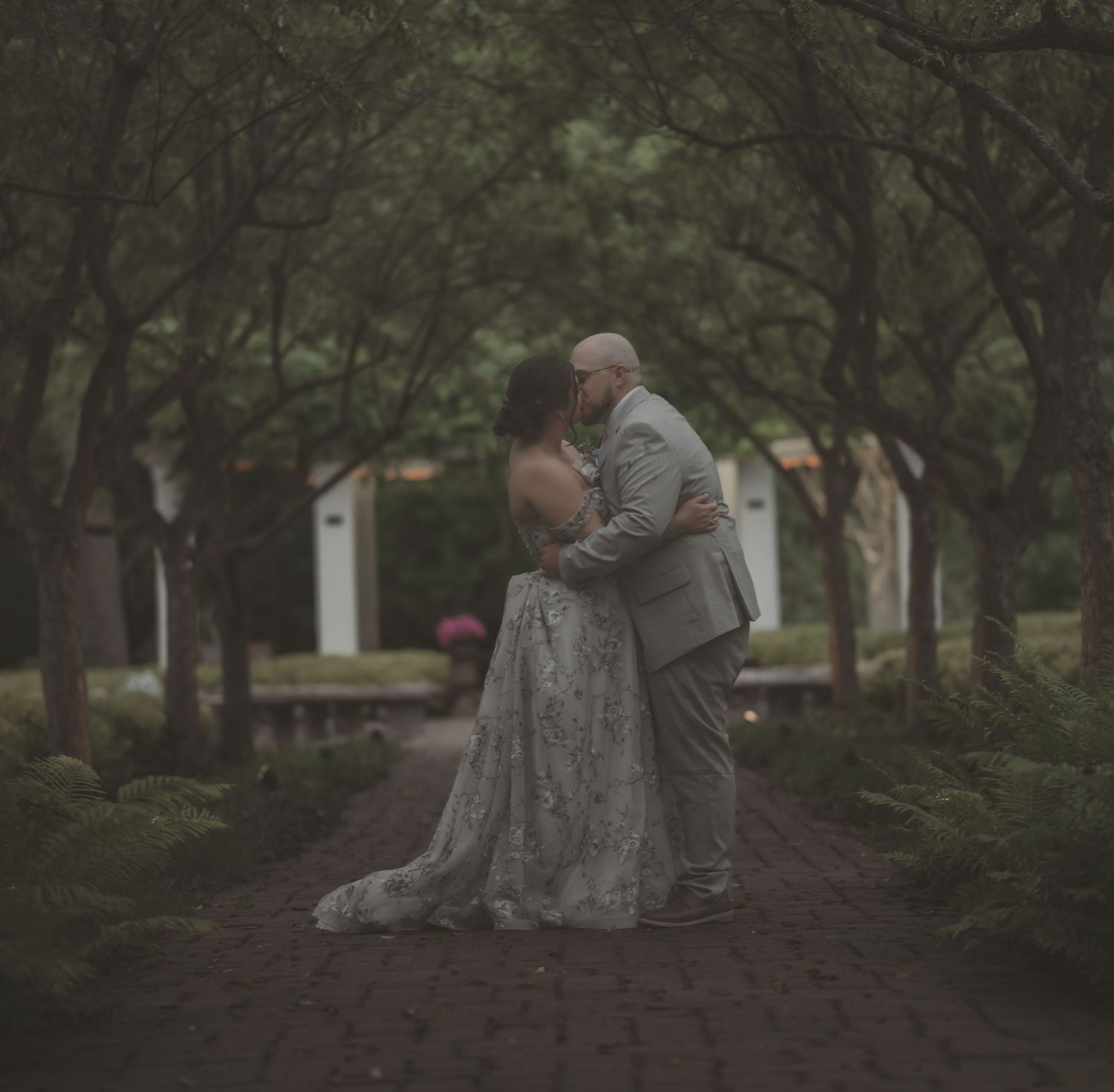 A couple shares an intimate embrace on a tree-lined garden path during their romantic wedding photoshoot.