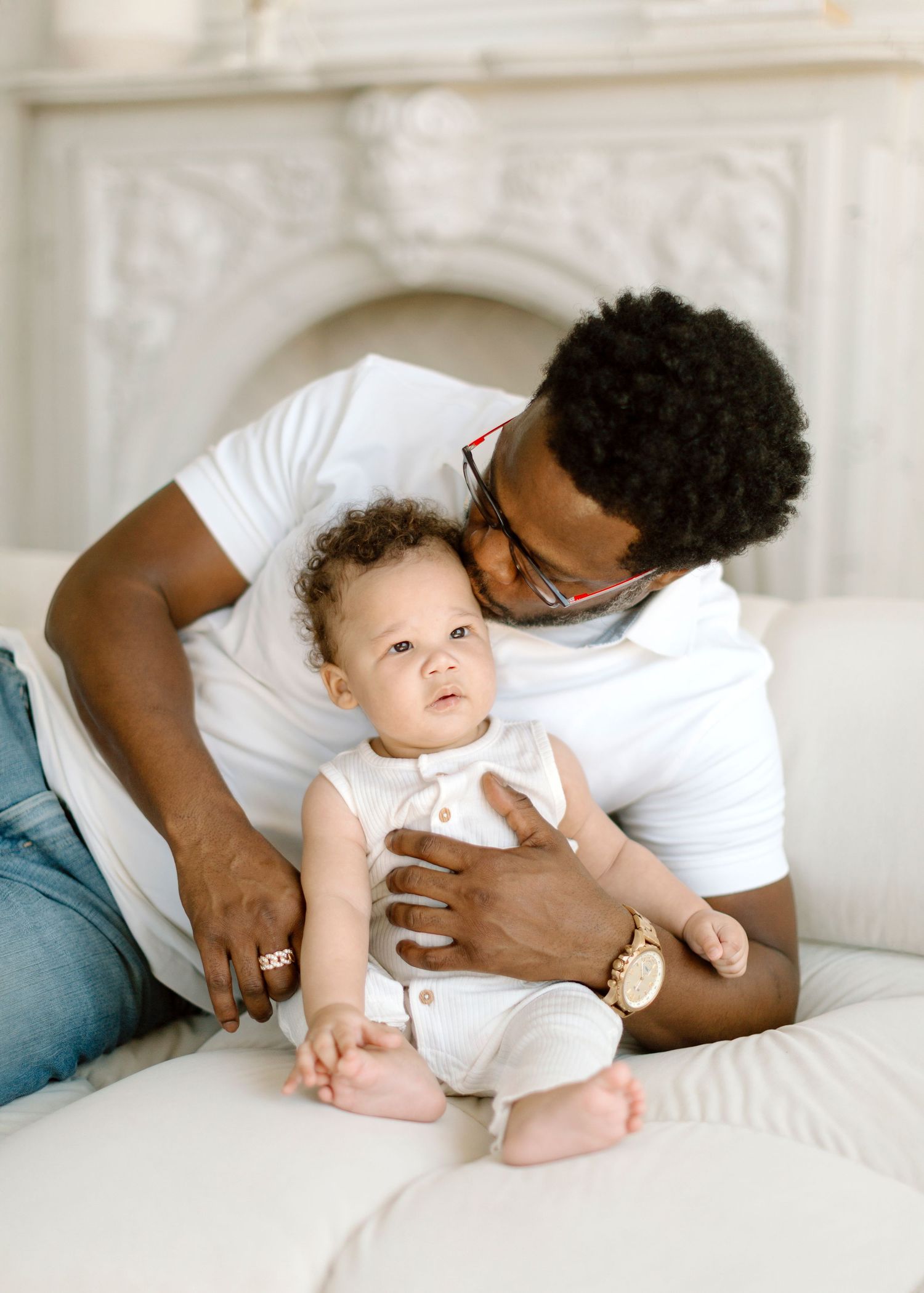 A baby wearing a white sleeveless onesie lies on cream-colored bedding while looking upward with a gentle expression.